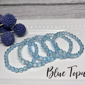 May include: Four blue topaz gemstone bracelets stacked on a white plate. The bracelets are made of round beads and have a light blue color. The text "Blue Topaz" is written in black cursive on the plate.