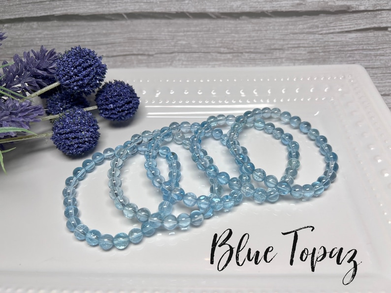 May include: Four blue topaz gemstone bracelets stacked on a white plate. The bracelets are made of round beads and have a light blue color. The text "Blue Topaz" is written in black cursive on the plate.