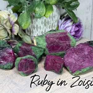 May include: Several raw ruby in zoisite stones with a mix of deep pink and green hues are displayed. The stones are arranged on a light-colored wooden surface, with the text "Ruby in Zoisite" in a handwritten font. The background includes flowers and greenery.