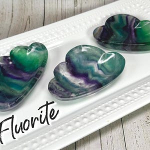 May include: Three fluorite cloud-shaped dishes in shades of green and purple sit on a white rectangular tray. The word "Fluorite" is written in a black script font across the bottom of the image.