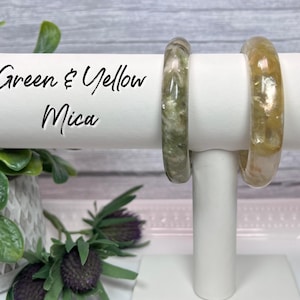 May include: Two resin bangles with green and yellow mica flecks. The bangles are displayed on a white stand. Green & Yellow Mica.