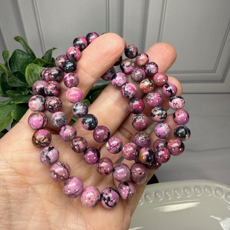 May include: A close-up of several beaded bracelets. The bracelets are made of round, pink and black stones. The bracelets are held in a hand, with a white plate and green plant in the background.