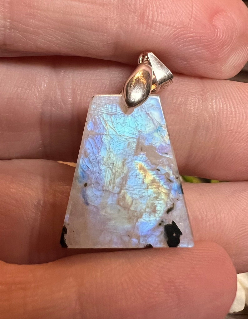 May include: A silver pendant with a large, iridescent, triangular moonstone. The stone has a white base with blue, green, and gold iridescence.