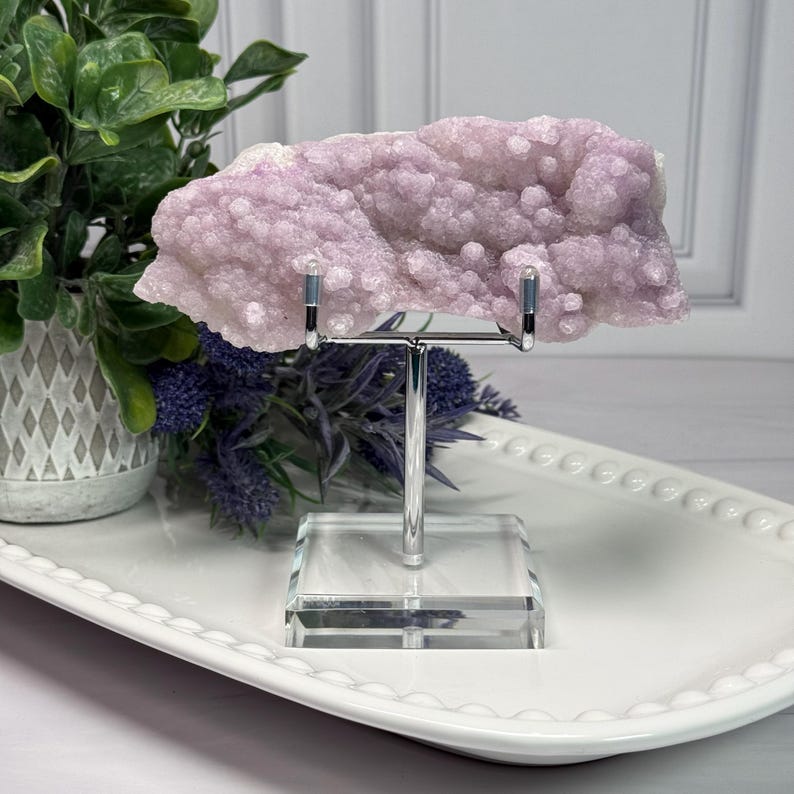 May include: A light purple crystal cluster displayed on a silver metal stand with a clear acrylic base. The crystal has a bubbly texture and is set against a white background. The stand is on a white, oval-shaped tray.