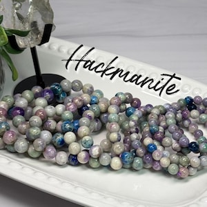 May include: A collection of beaded bracelets made from round, polished Hackmanite stones in shades of purple, blue, white, and grey. The bracelets are arranged on a white, rectangular, beaded-edge tray with the word Hackmanite written in black.