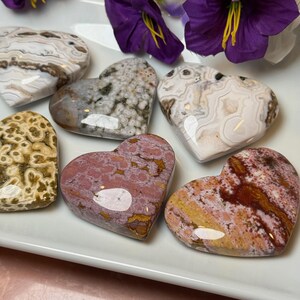 May include: A collection of heart-shaped stones in various colors and patterns, including white, brown, pink, and yellow. The stones are arranged on a white surface, with purple flowers in the background. These decorative stones are ideal for home decor.