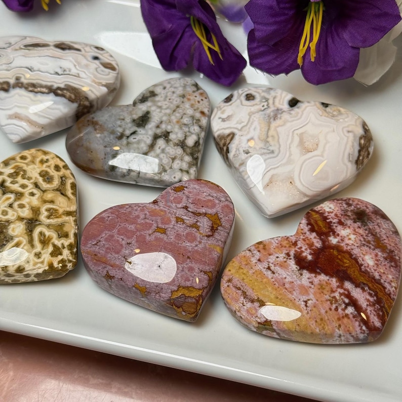 May include: A collection of heart-shaped stones in various colors and patterns, including white, brown, pink, and yellow. The stones are arranged on a white surface, with purple flowers in the background. These decorative stones are ideal for home decor.