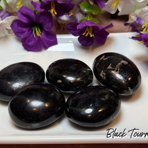 May include: Five polished black tourmaline stones on a white surface, with purple and white flowers in the background. The stones are oval-shaped and have a glossy finish. The text "Black Tourmaline" is written in a script font.
