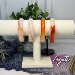 May include: Four agate bracelets in shades of orange, white, and pink are displayed on a white stand. The bracelets have a polished, smooth surface. The word "Agate" is written in cursive on the bottom right.