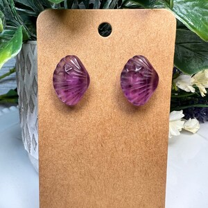 Op de afbeelding: Een paar paarse schelpvormige oorbellen met een swirl-patroon. De oorbellen worden gepresenteerd op een bruine kaart.