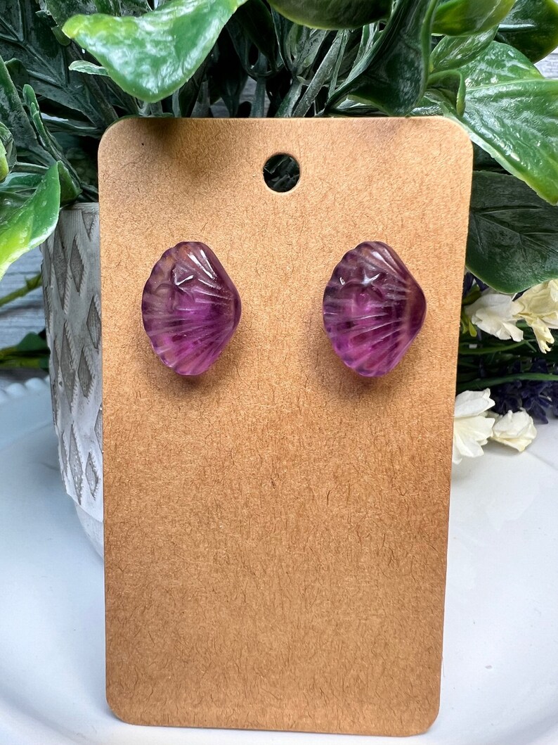 Op de afbeelding: Een paar paarse schelpvormige oorbellen met een swirl-patroon. De oorbellen worden gepresenteerd op een bruine kaart.