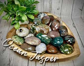 May include: A wooden bowl filled with polished Ocean Jasper stones in various colors, including green, brown, and white. The stones are smooth and oval-shaped. The words "Ocean Jasper" are written in white script across the bowl.
