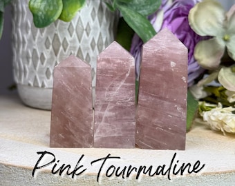 May include: Three polished pink tourmaline crystal towers of varying heights standing upright. Each stone features a translucent, dusty rose hue with natural internal veining and a pointed, faceted top. The text Pink Tourmaline is visible.