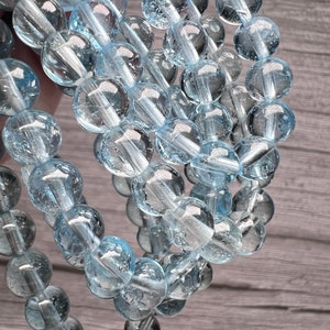 May include: A strand of light blue aquamarine beads, each bead is round and has a smooth, polished surface. The beads are strung together on a clear thread.