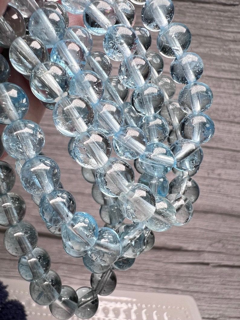 May include: A strand of light blue aquamarine beads, each bead is round and has a smooth, polished surface. The beads are strung together on a clear thread.