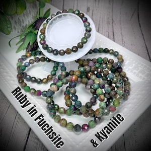 May include: A collection of gemstone bracelets featuring ruby in fuchsite and kyanite. The bracelets are made of faceted, round beads in shades of green, brown, purple, and red. One bracelet rests on a white, circular display stand. The text "Ruby in Fuchsite & Kyanite" is visible.