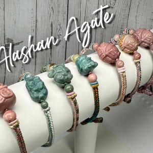 May include: A collection of Alashan agate bracelets. Each bracelet features a carved stone charm in shades of pink and green, strung on a braided cord. The charms depict various figures, and the cords have colorful accents.