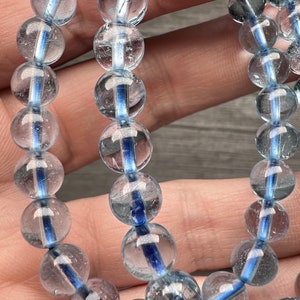 May include: Close-up of two aquamarine bracelets. Each bracelet features clear, round beads with a vibrant blue stripe through the centre. The beads are strung together, creating a delicate and elegant accessory. The bracelets are displayed against a neutral background.