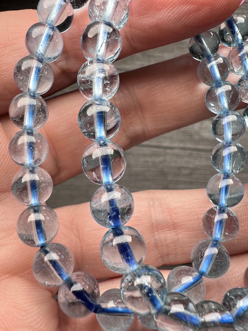 May include: Close-up of two aquamarine bracelets. Each bracelet features clear, round beads with a vibrant blue stripe through the centre. The beads are strung together, creating a delicate and elegant accessory. The bracelets are displayed against a neutral background.