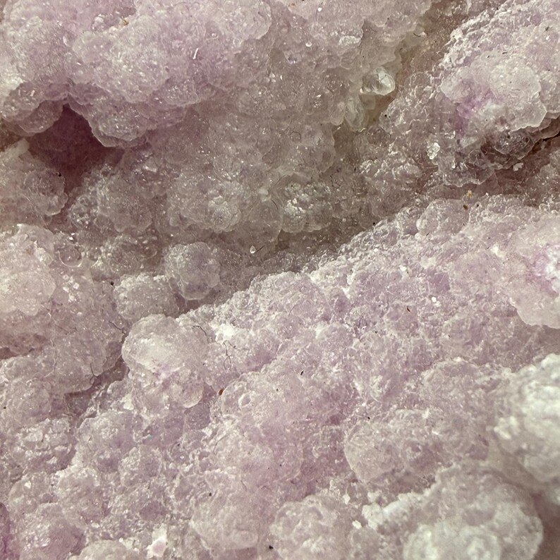 May include: Close-up of a crystalline formation with a soft, textured appearance. The surface is a pale lavender color, with rounded, clustered shapes. The overall effect is delicate and organic, suggesting a mineral or geological specimen.