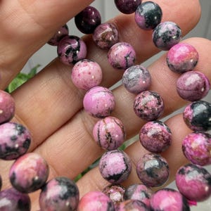 May include: A close-up of a bracelet made of round, polished beads. The beads are primarily pink with dark gray and black marbling. The bracelet is held in a hand, with the beads appearing to be approximately 0.4 inches in diameter.