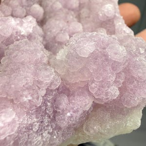 May include: Close-up of a cluster of light purple crystals. The crystals have a bubbly, textured appearance, with a mix of light and darker shades of purple. The overall shape is irregular and organic.