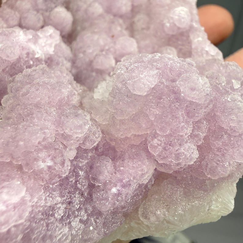 May include: Close-up of a cluster of light purple crystals. The crystals have a bubbly, textured appearance, with a mix of light and darker shades of purple. The overall shape is irregular and organic.