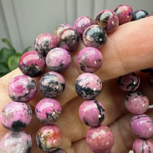 May include: Close-up of a beaded bracelet featuring round, polished beads. The beads are primarily pink with black and gray marbling. The bracelet is held by a person's fingers, showcasing the texture and color variations of the gemstones.