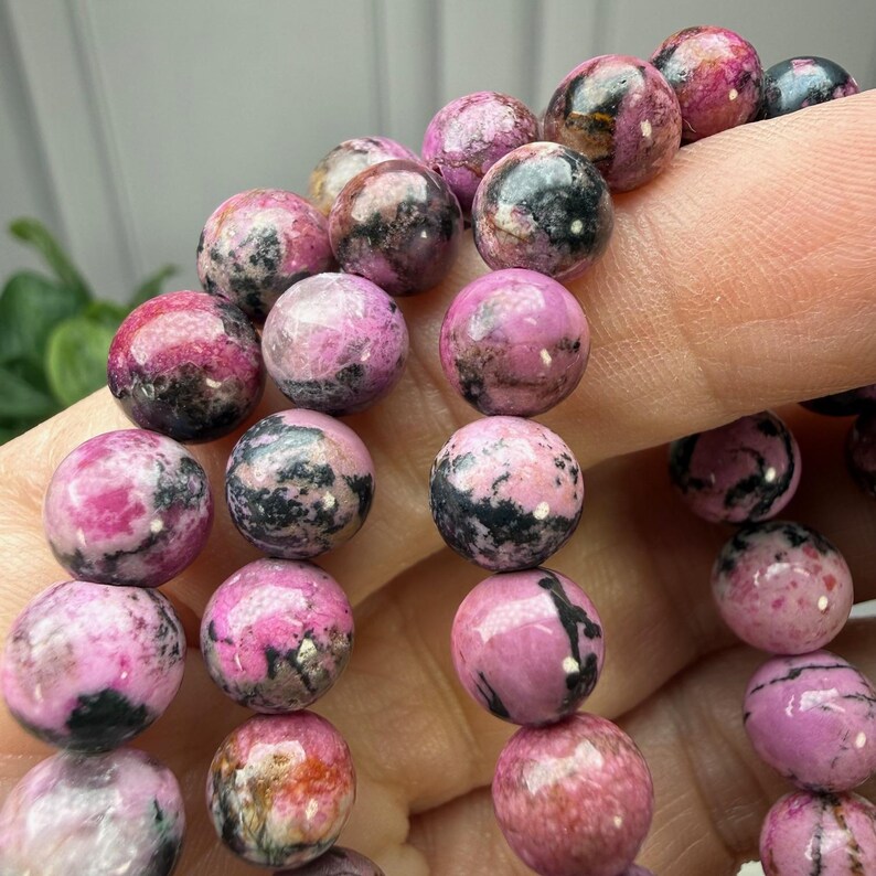 May include: Close-up of a beaded bracelet featuring round, polished beads. The beads are primarily pink with black and gray marbling. The bracelet is held by a person's fingers, showcasing the texture and color variations of the gemstones.