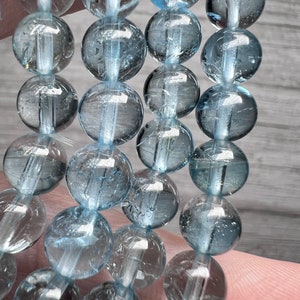 May include: A strand of light blue aquamarine beads. The beads are round and have a smooth, polished finish. The beads are strung on a clear elastic cord.
