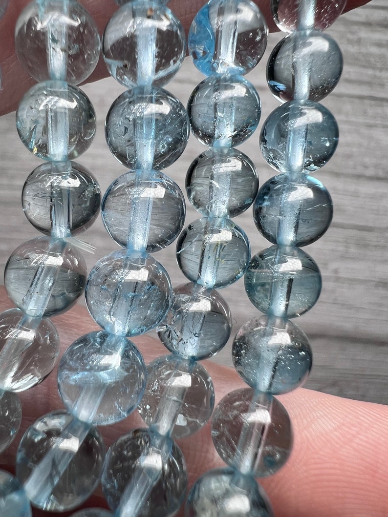 May include: A strand of light blue aquamarine beads. The beads are round and have a smooth, polished finish. The beads are strung on a clear elastic cord.