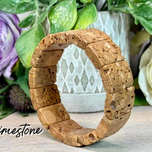 May include: A light brown limestone bracelet with a rectangular block design. The bracelet is displayed in front of a blurred background of flowers and greenery. The word "Limestone" is written in cursive.