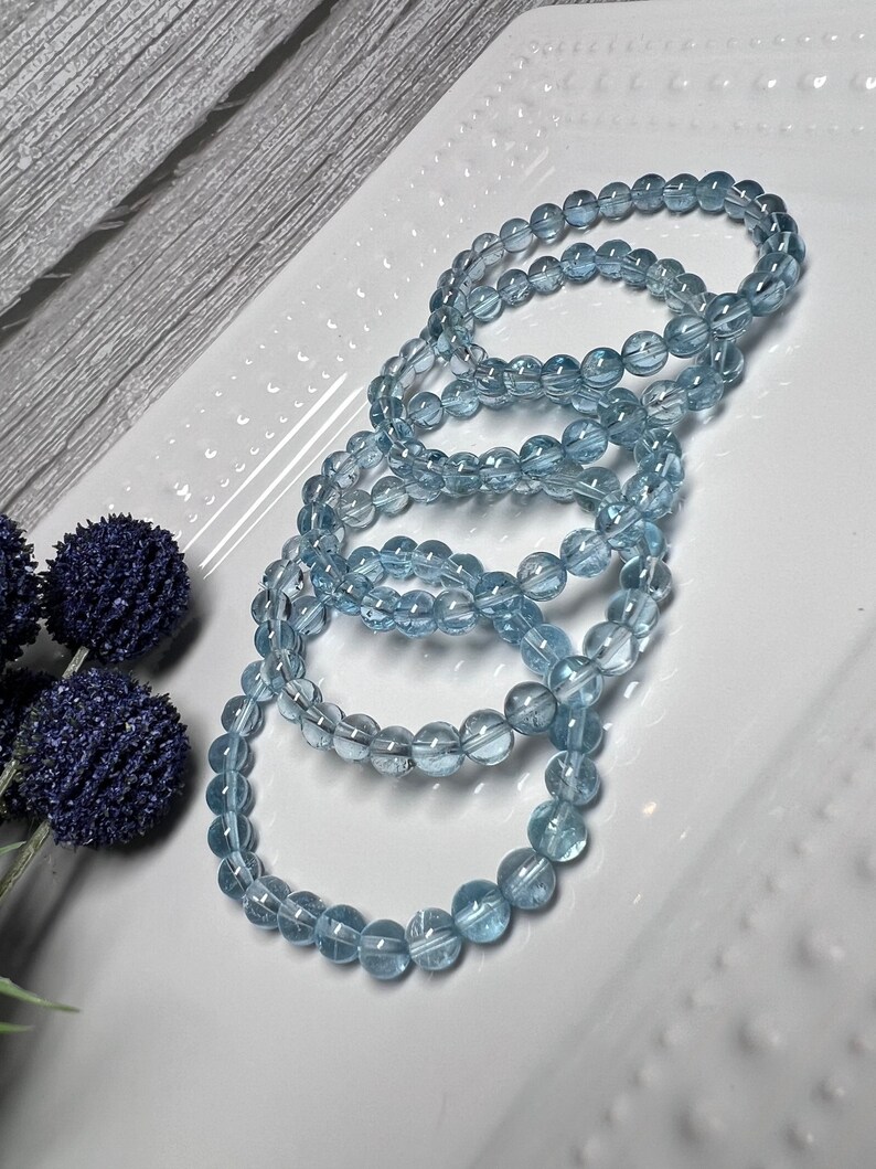 May include: Four light blue aquamarine gemstone bracelets stacked on a white plate. The bracelets are made of round beads.