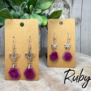 May include: Two pairs of earrings displayed on brown cards. One pair features silver-toned lotus flower charms, and the other has butterfly charms. Both pairs have vibrant, irregular-shaped, deep pink ruby stones. The word "Ruby" is written in cursive.