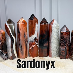 May include: Eight polished sardonyx crystal towers arranged in a row, each featuring distinct banded patterns in shades of deep red, brown, black, and white, with some pieces displaying translucent crystalline textures and the text SARDONYX.
