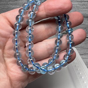 May include: Two bracelets composed of clear, round beads with a light blue interior. The bracelets are held in a hand, highlighting the translucent quality of the beads. The background is a wooden surface and a white dish.