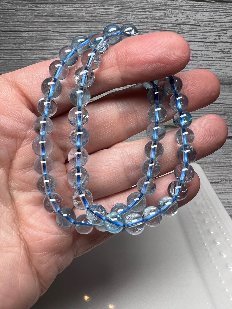 May include: Two bracelets composed of clear, round beads with a light blue interior. The bracelets are held in a hand, highlighting the translucent quality of the beads. The background is a wooden surface and a white dish.