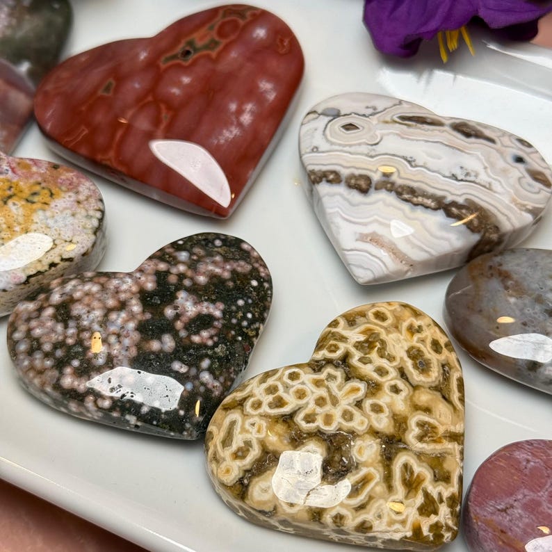 May include: A collection of polished heart-shaped stones in various colors and patterns. The stones include shades of red, brown, white, and green, with unique veining and speckled textures. The hearts are arranged on a white surface.
