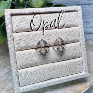 May include: Two silver rings with opal stones set in a white fabric ring display box. The box has the word "Opal" written on the top.
