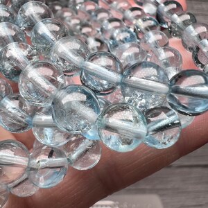 May include: A strand of light blue aquamarine beads, each bead is round and smooth. The beads are strung on a clear thread and are arranged in a staggered pattern.