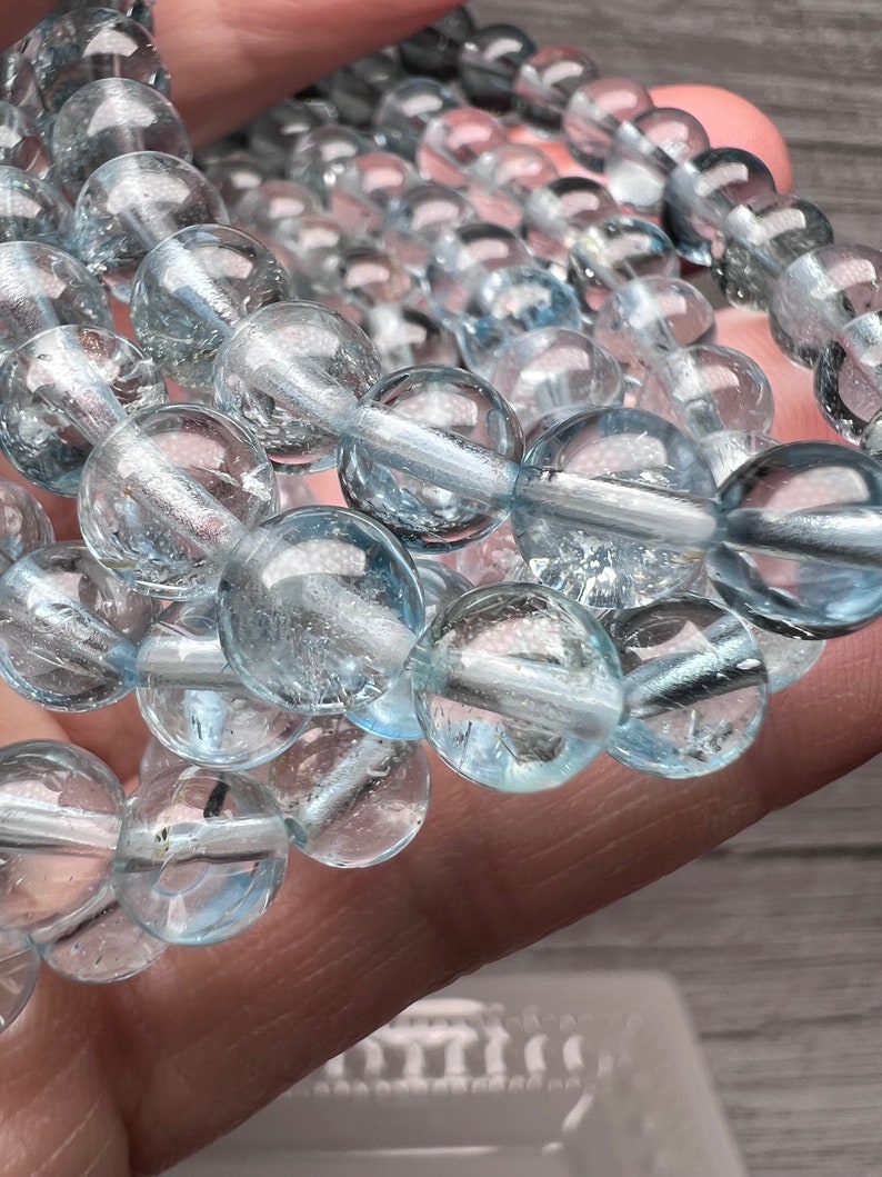 May include: A strand of light blue aquamarine beads, each bead is round and smooth. The beads are strung on a clear thread and are arranged in a staggered pattern.