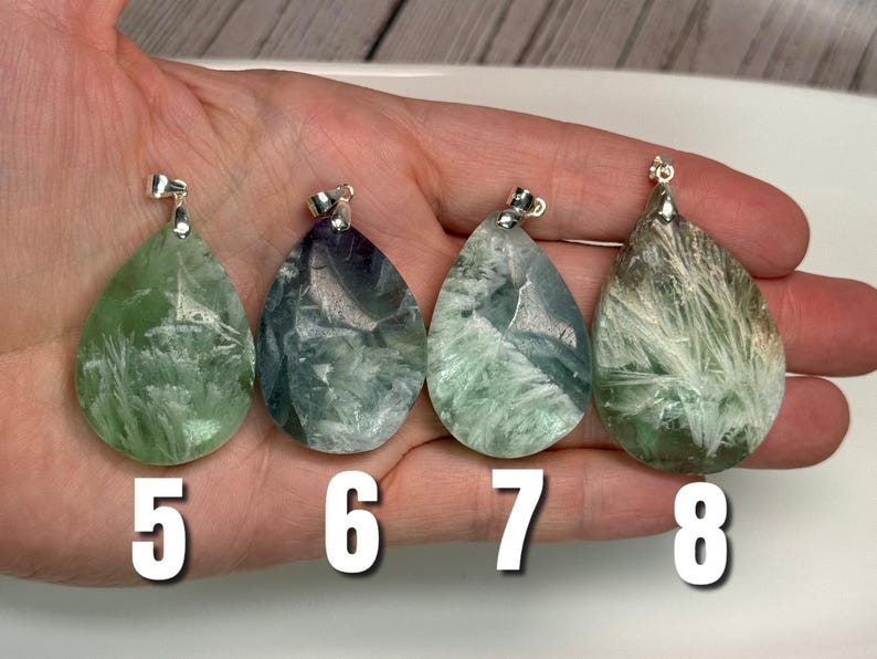 May include: Four teardrop-shaped fluorite pendants with silver bails. The pendants display green, blue, and purple hues with crystalline patterns. Numbers 5, 6, 7, and 8 are printed in white below the pendants.