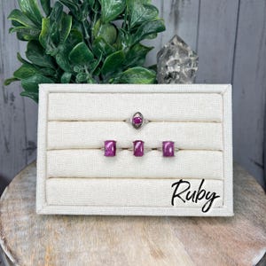 May include: Four silver rings with pink ruby gemstones displayed on a white fabric-covered ring display. The rings are arranged in a row, with the top ring featuring an oval-shaped stone and the three bottom rings featuring square-shaped stones. The display is labeled "Ruby".