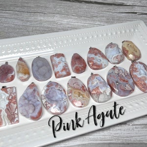 May include: A collection of pink agate pendants with various shapes and patterns. The pendants are arranged on a white plate with a decorative border. The text "Pink Agate" is written in black below the pendants.