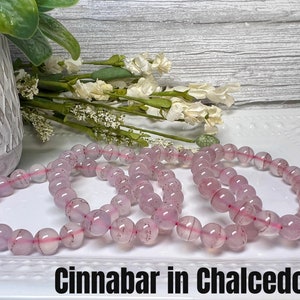May include: Three pink chalcedony bracelets with red cinnabar inclusions. The bracelets are stacked on a white surface. The text "cinnabar in chalcedony" is written below the bracelets.