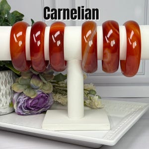 May include: Six smooth, polished carnelian bangles displayed on a white T-bar stand. The bracelets feature varying shades of translucent orange, red, and brown with natural banding patterns. The word Carnelian is printed in black above the stand.