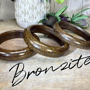 May include: Three brown and gold bronzite bracelets are displayed on a wooden surface. The bracelets are oval-shaped and have a polished finish. The word "Bronzite" is written in a cursive font.