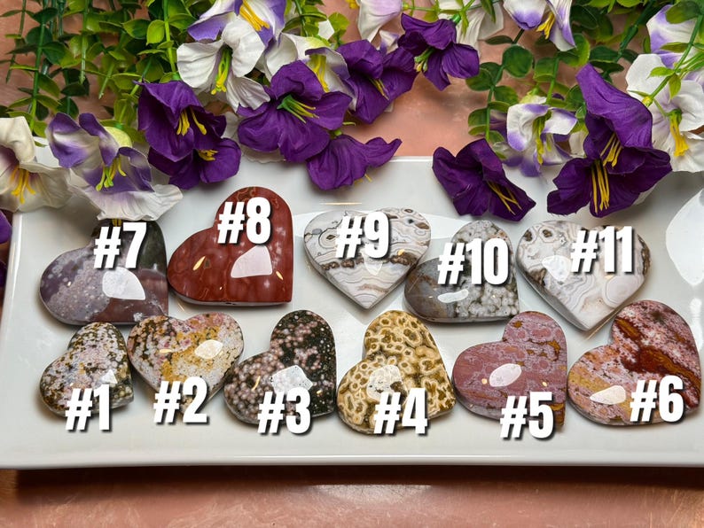 May include: Eleven heart-shaped stones in various colors and patterns are displayed on a white tray, each labeled with a number. The stones range from speckled to solid, with colors including red, brown, and gray. Purple and white flowers are in the background.