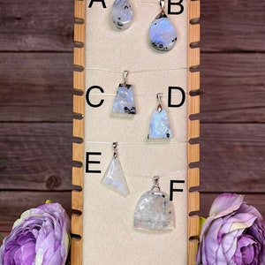 May include: Six rainbow moonstone pendants on display on a wooden stand. The pendants are labeled A through F.