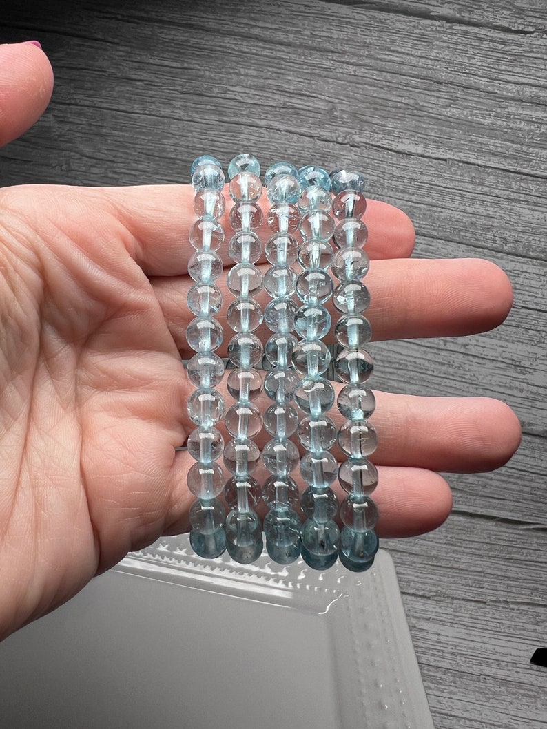 May include: Five strands of light blue aquamarine bead bracelets. The beads are round and have a translucent appearance.